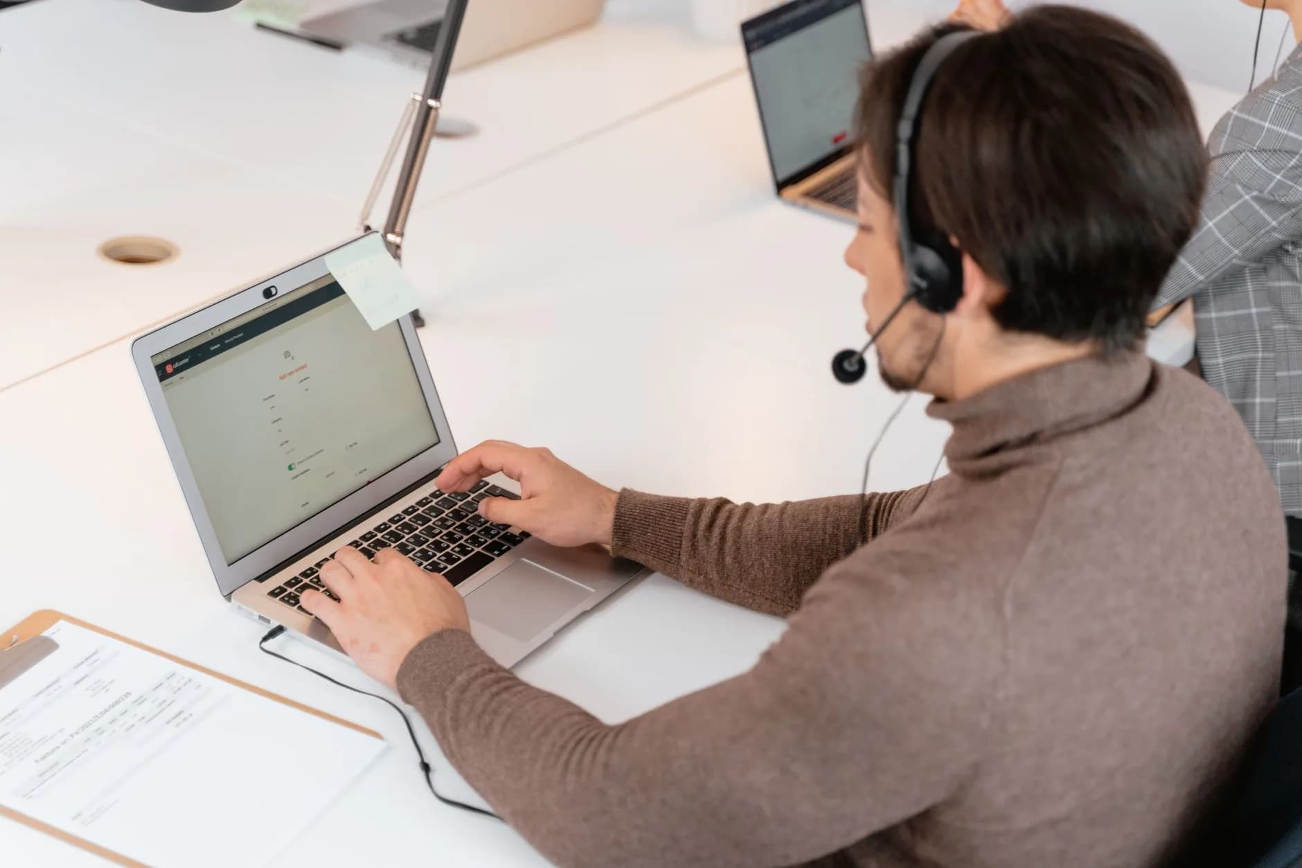 Agente di call center con cuffie che lavora su un laptop in un ambiente d'ufficio moderno.