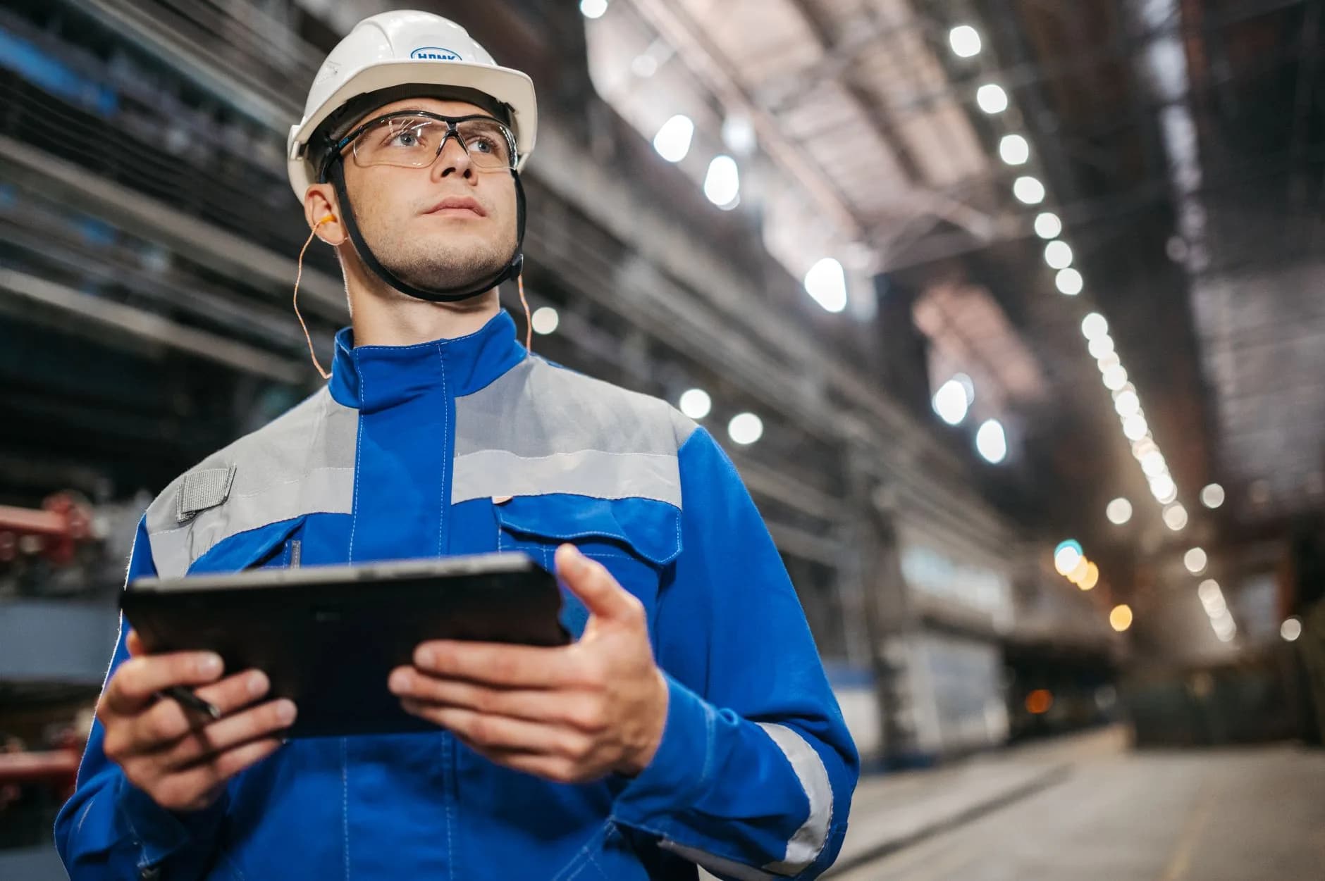 Operaio in tuta di sicurezza che utilizza un tablet in un ambiente industriale ben illuminato.